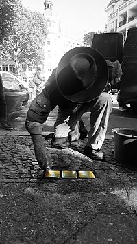 Gunter Demnig beim Verlegen von STOLPERSTEINEN. Schwarzweiß-Foto mit messingfarben hervorgehobenen STOLPERSTEINEN